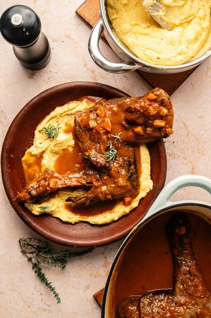 A plate of Mediterranean Lamb Shank with rich brown sauce is served over creamy polenta, with a pot of soft polenta and a Dutch oven of tender lamb shank nearby.