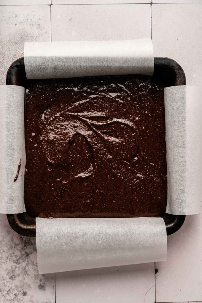 A square baking pan lined with parchment paper is filled with rich Protein Brownies batter, ready to be baked.