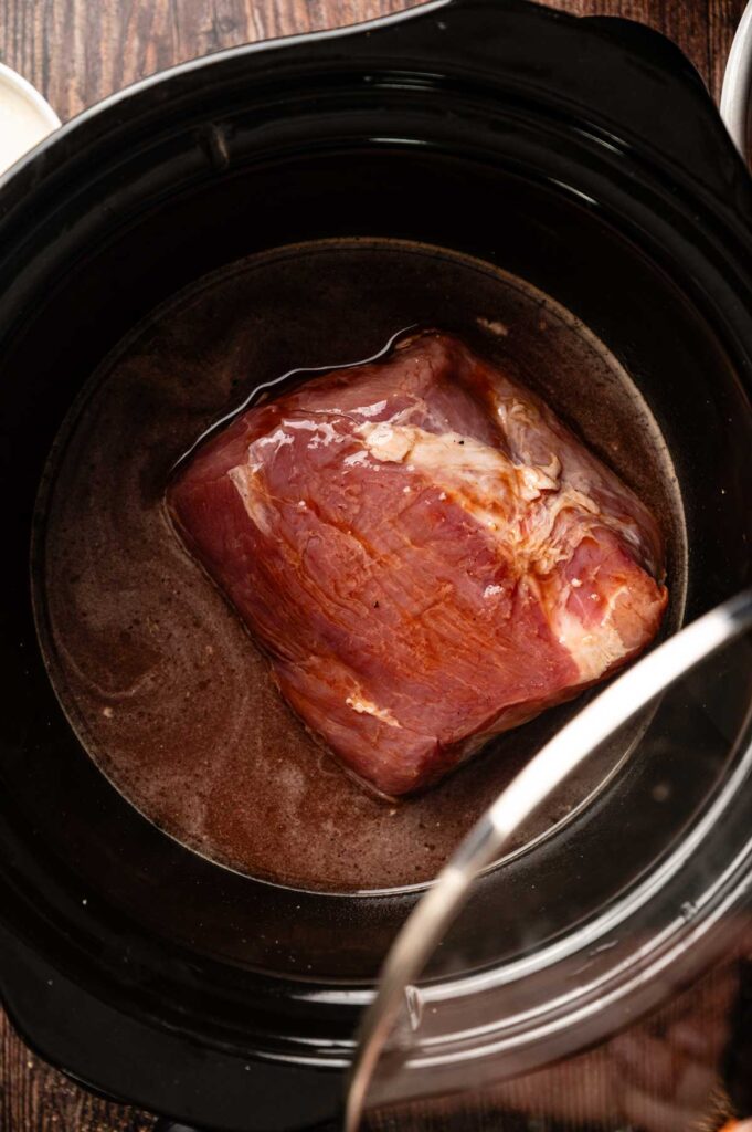A piece of raw meat sits in liquid inside a black slow cooker on a wooden surface, ready to be transformed into tender pulled pork sliders.