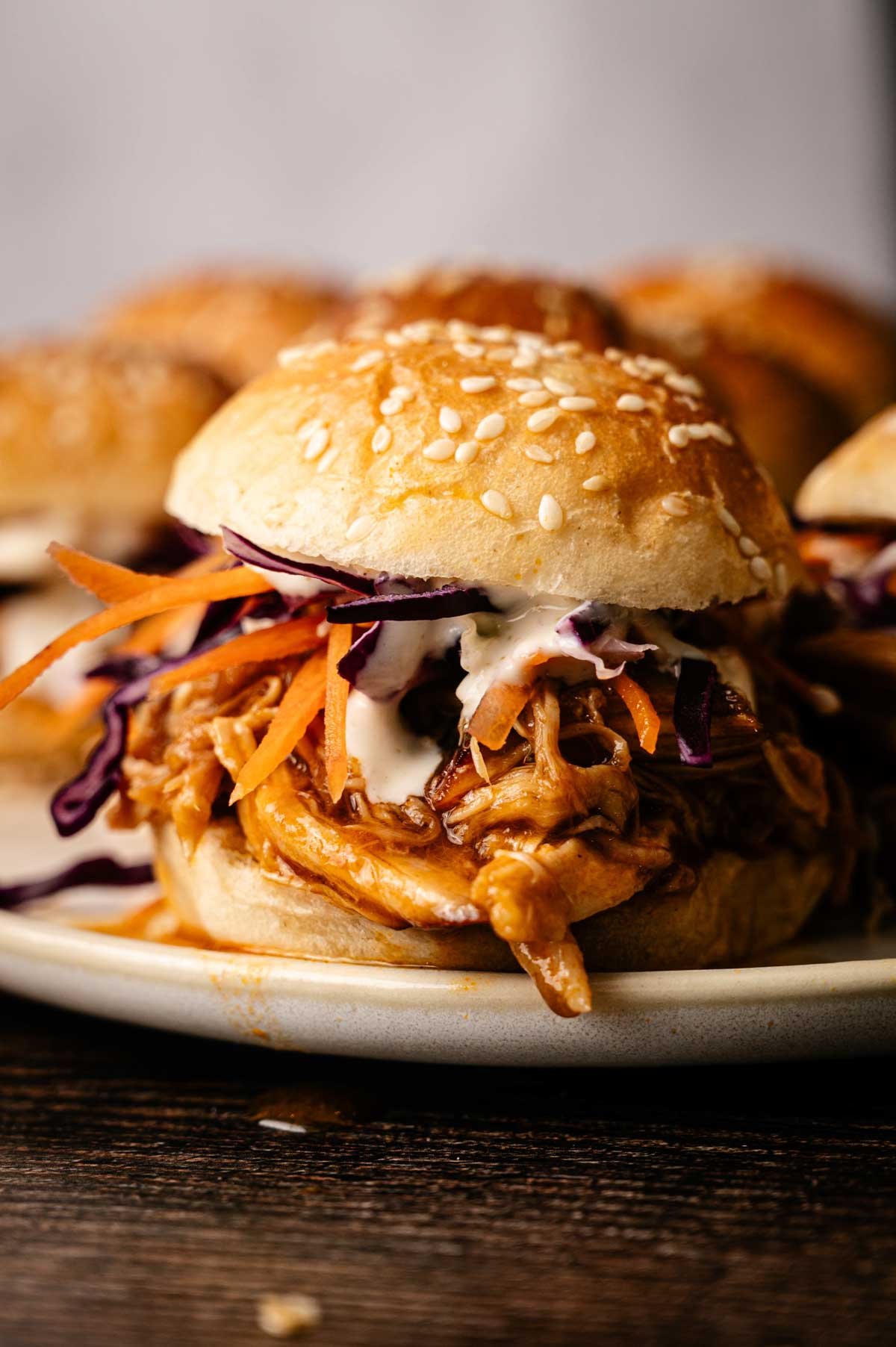 A pulled chicken sandwich with barbecue sauce, shredded carrots, purple cabbage, and creamy dressing on a sesame bun, served on a plate—perfect alongside savory Pulled Pork Sliders for a delicious meal.
