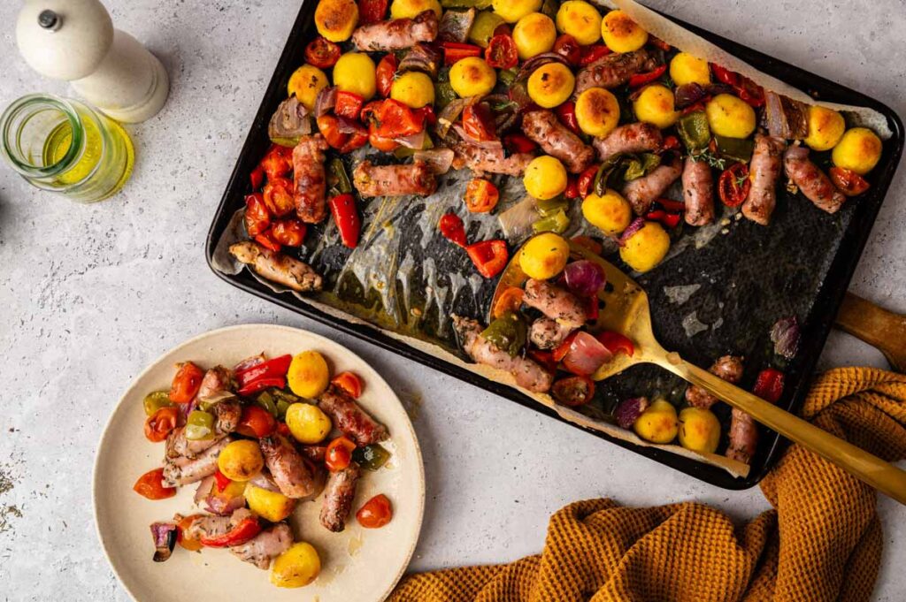 Sheet Pan Gnocchi with roasted sausages, cherry tomatoes, bell peppers, and red onions is served on a plate with a yellow cloth beside it.