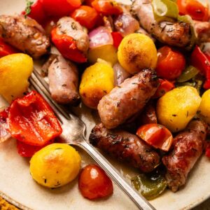 A plate of roasted sausages, potatoes, cherry tomatoes, red onions, bell peppers, and Sheet Pan Gnocchi sits ready to enjoy, with a fork on the side.
