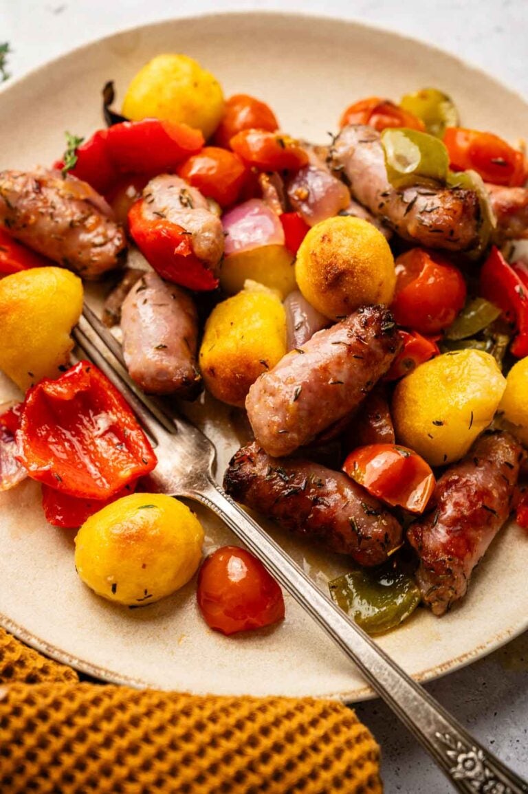 A plate of roasted sausages, potatoes, cherry tomatoes, red onions, bell peppers, and Sheet Pan Gnocchi sits ready to enjoy, with a fork on the side.