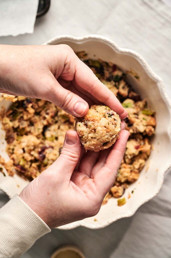 Two hands are forming a ball of Air Fryer Stuffing Balls mixture over a bowl filled with more stuffing on a light-colored surface.