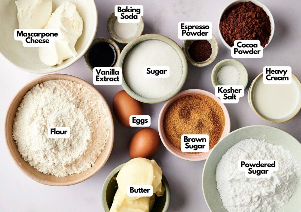 Bowls and plates containing ingredients for Air Fryer Tiramisu Cookies—mascarpone cheese, flour, butter, eggs, sugars, baking soda, cocoa powder, espresso powder, vanilla extract, kosher salt, and heavy cream—are arranged on a counter.