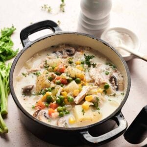 A black pot filled with creamy Air Fryer Chicken Pot Pie Soup, brimming with potatoes, peas, corn, carrots, and mushrooms, sits on a light surface near celery, a pepper grinder, and a salt bowl.