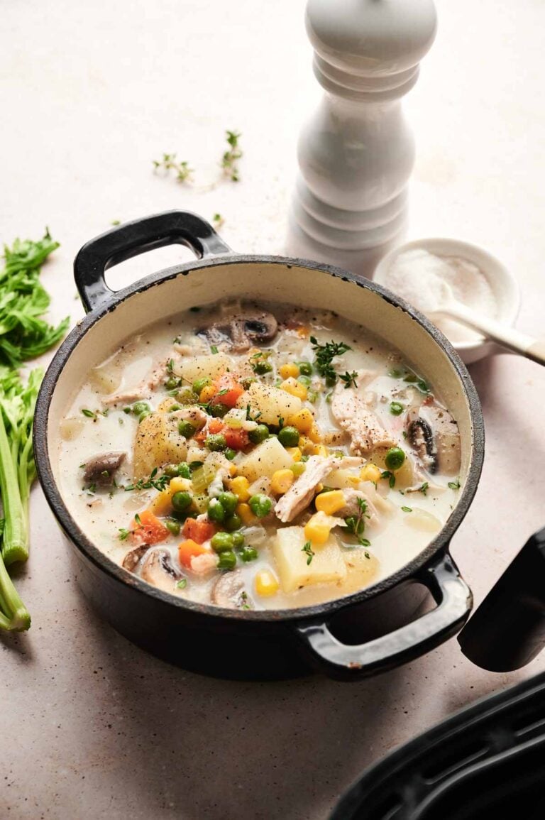 A black pot filled with creamy Air Fryer Chicken Pot Pie Soup, brimming with potatoes, peas, corn, carrots, and mushrooms, sits on a light surface near celery, a pepper grinder, and a salt bowl.