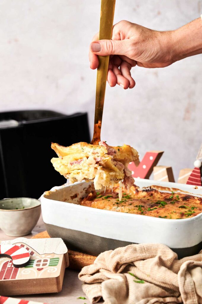 A hand uses a gold utensil to serve cheesy baked pasta from a rectangular white casserole dish on a table with holiday decorations, creating a festive scene reminiscent of classics like Air Fryer Scalloped Potatoes and Ham.
