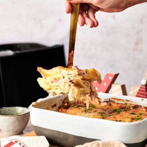 A hand serves a portion of baked pasta casserole from a ceramic dish, with holiday decorations and a cloth napkin in the foreground—perfect alongside Air Fryer Scalloped Potatoes and Ham for your festive table.