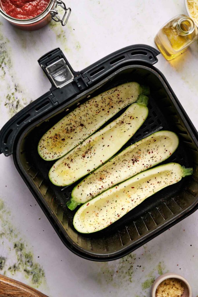 Four halved zucchinis seasoned with pepper and herbs are placed inside an air fryer basket, ready to be cooked into delicious Air Fryer Zucchini Boats.