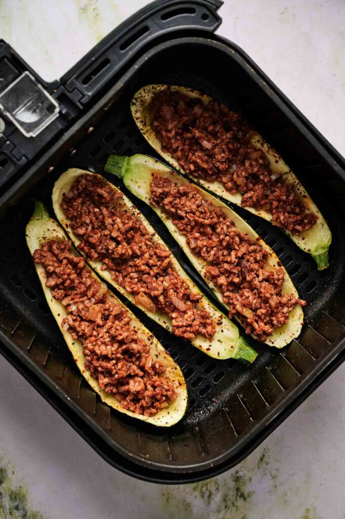 Four Air Fryer Zucchini Boats, each brimming with seasoned ground meat, are placed inside an air fryer basket, ready to be cooked.