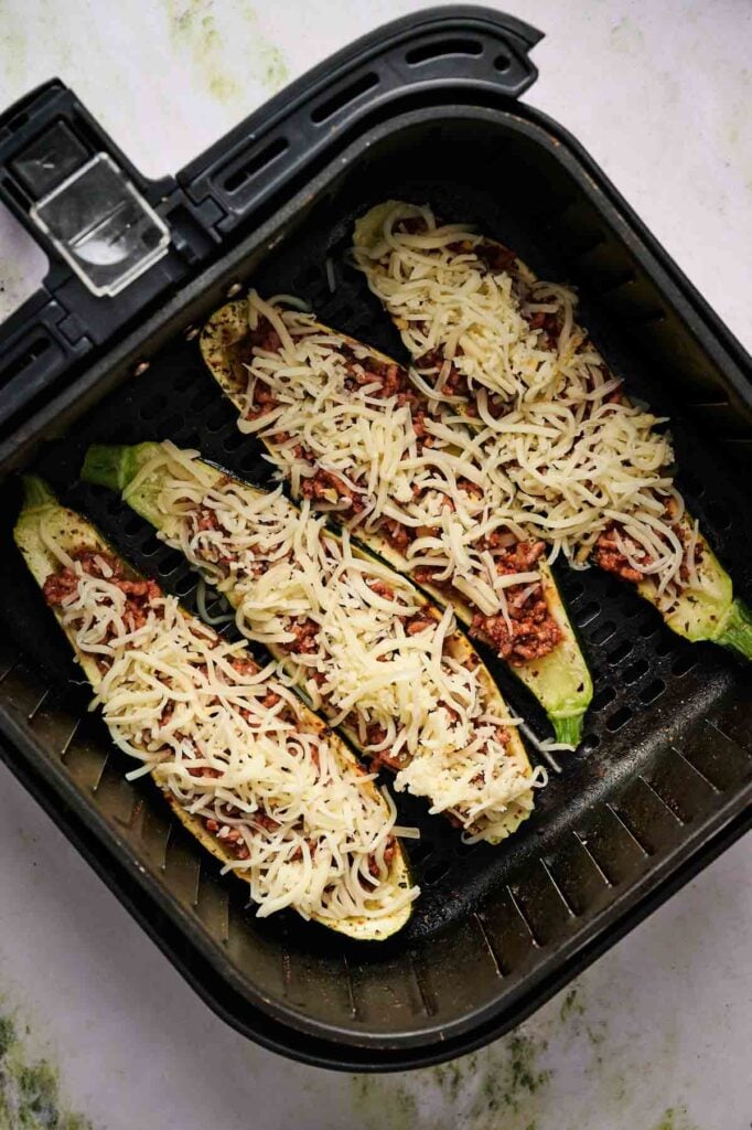 Four Air Fryer Zucchini Boats, topped with savory meat sauce and melted cheese, are arranged in the basket, ready to be cooked to perfection.