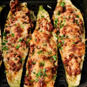Three Air Fryer Zucchini Boats filled with ground meat and melted cheese, garnished with chopped herbs, inside an air fryer basket.