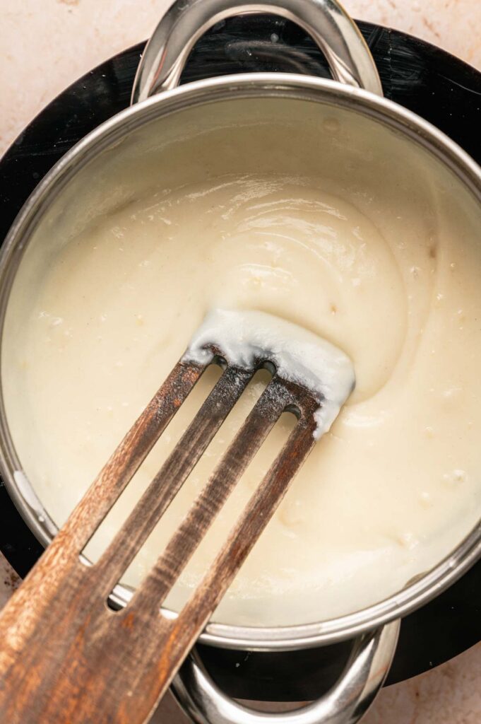 A stainless steel pot containing a thick white sauce for Chicken Croquettes is being stirred with a wooden slotted spatula.