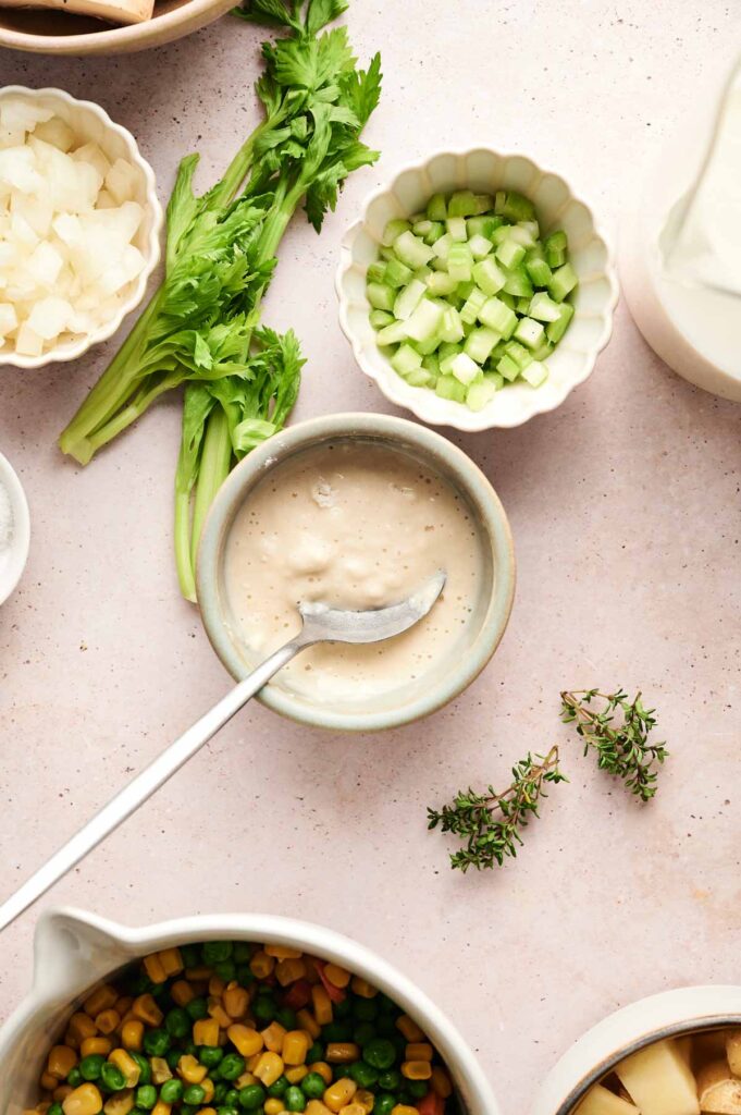 Bowl of creamy sauce with spoon, surrounded by chopped celery, onions, mixed vegetables, potato cubes, fresh herbs, and a pitcher of liquid on a light surface—perfect ingredients for an Air Fryer Chicken Pot Pie Soup.