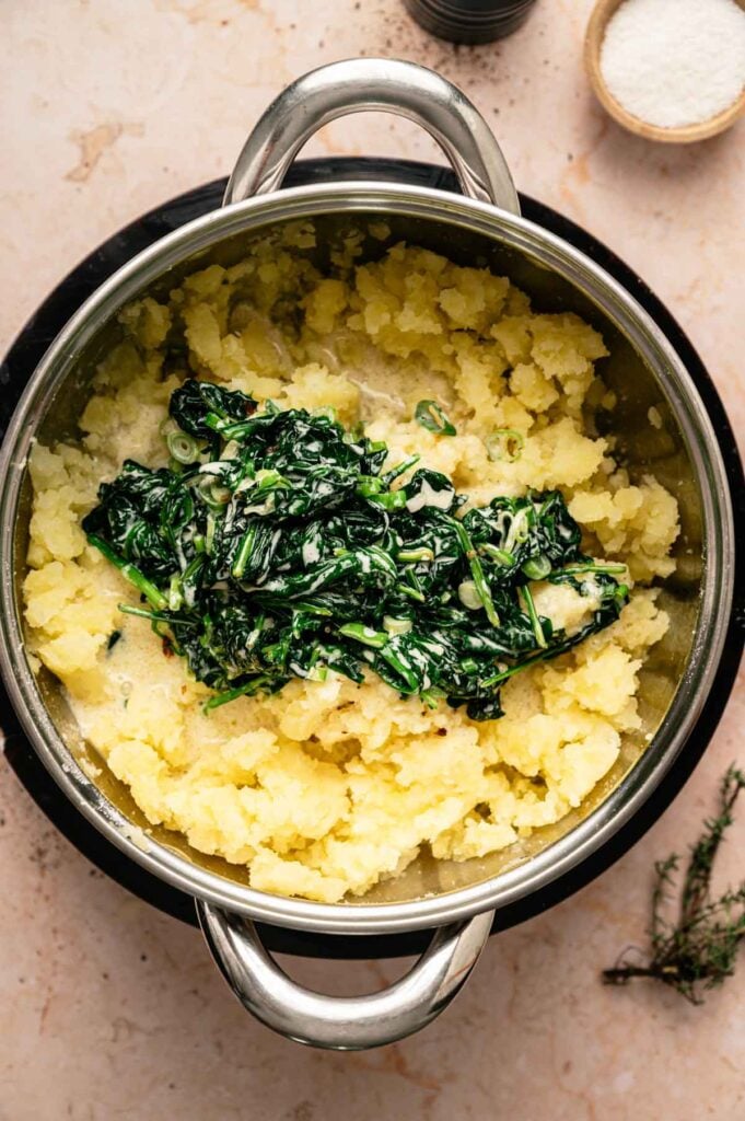 A pot of creamy colcannon—mashed potatoes blended with cooked spinach and fresh herbs—sits on a light countertop, flanked by small bowls of salt and spices.