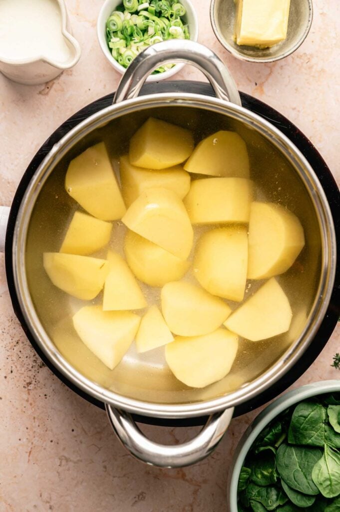 A pot of peeled potato chunks in water, surrounded by a bowl of spinach, sliced green onions, a block of butter, and a pitcher of cream on a countertop—everything you need to prepare traditional Colcannon.