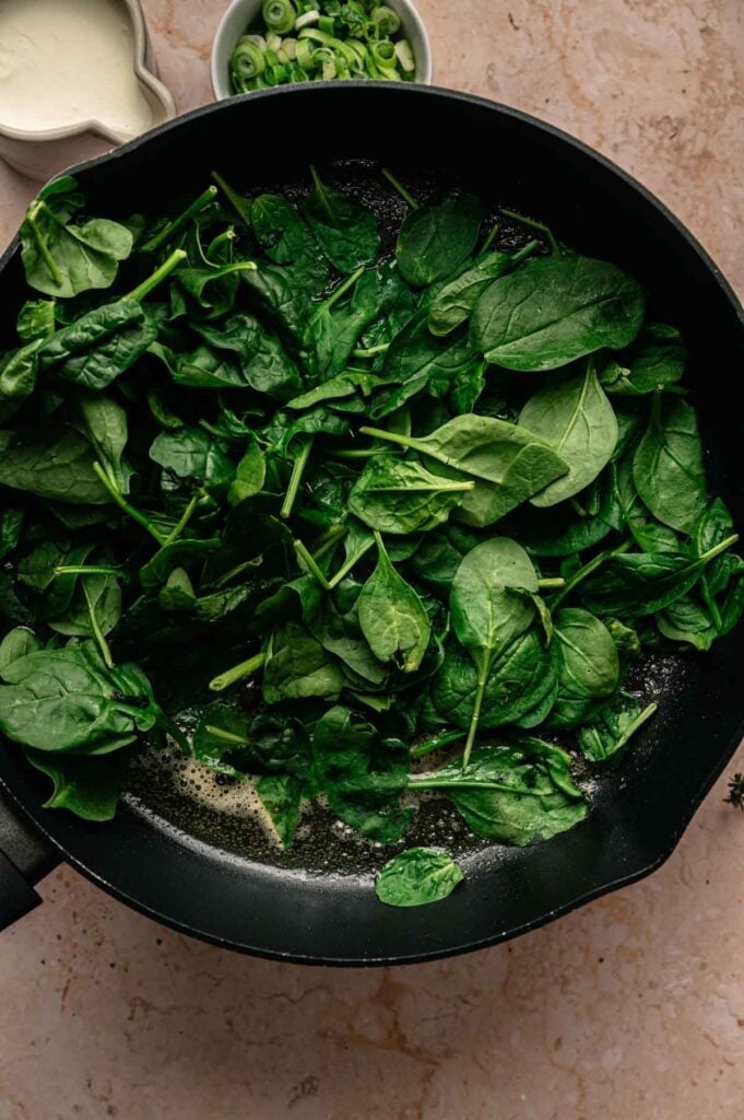 Fresh spinach leaves in a black skillet, beginning to wilt in melted butter, evoke the comforting flavors of colcannon. In the background, bowls of sliced green onions and cream rest on a light countertop, ready to be added to this classic dish.