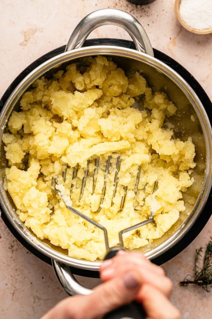 A hand mashes boiled potatoes in a stainless steel pot with a potato masher on a light-colored countertop, preparing the base for classic colcannon.