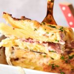 A close-up of Air Fryer Scalloped Potatoes and Ham shows layers of sliced potatoes, ham, creamy sauce, and a golden brown top, lifted by a fork from a baking dish.