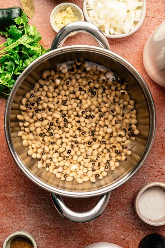 A pot of uncooked black-eyed peas, the essential base for Hoppin’ John, is surrounded by bowls of chopped onions, minced garlic, cilantro, spices, and seasonings on a brown surface.