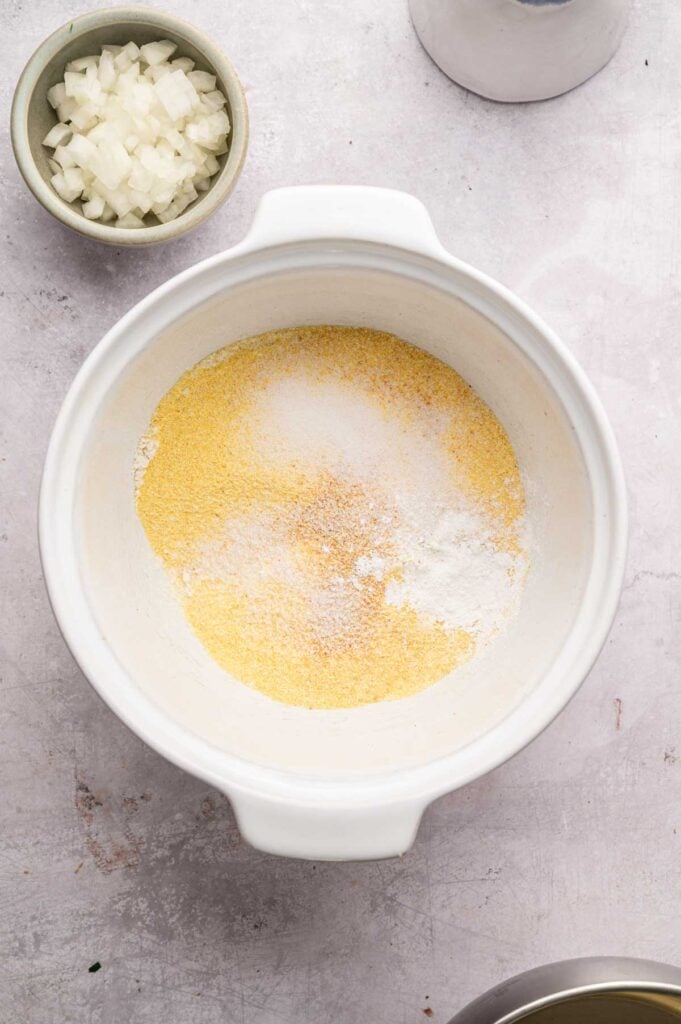 Cornmeal, flour, and spices in a white bowl next to a small dish of chopped onion