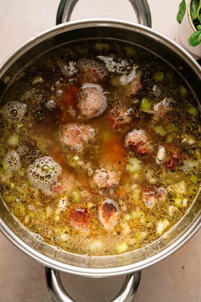 A pot of Italian Wedding Soup with meatballs, chopped celery, herbs, and broth simmering on a stovetop.