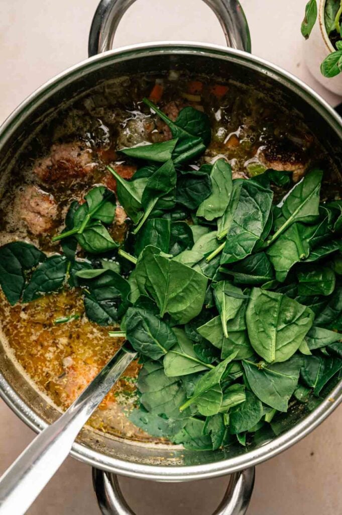 Large pot of Italian Wedding Soup with fresh spinach leaves on top, being stirred with a metal spoon.