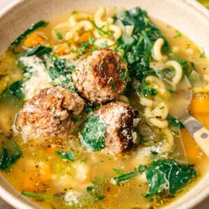 A bowl of Italian Wedding Soup with meatballs, spinach, macaroni pasta, and chunks of orange vegetables, garnished with grated cheese and herbs.