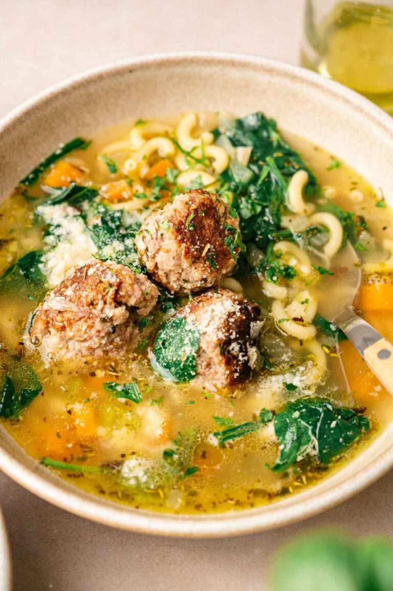 A bowl of Italian Wedding Soup with meatballs, spinach, macaroni pasta, and chunks of orange vegetables, garnished with grated cheese and herbs.