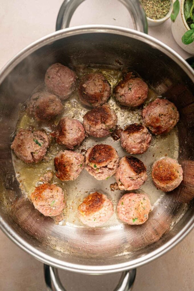 Several meatballs are browning in a stainless steel pot with oil, perfect for Italian Wedding Soup. Some herbs are visible in the background.
