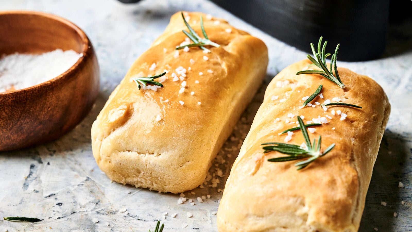 Two rectangular pieces of golden-brown bread topped with coarse salt and rosemary, placed on a light surface next to a small wooden bowl of salt.