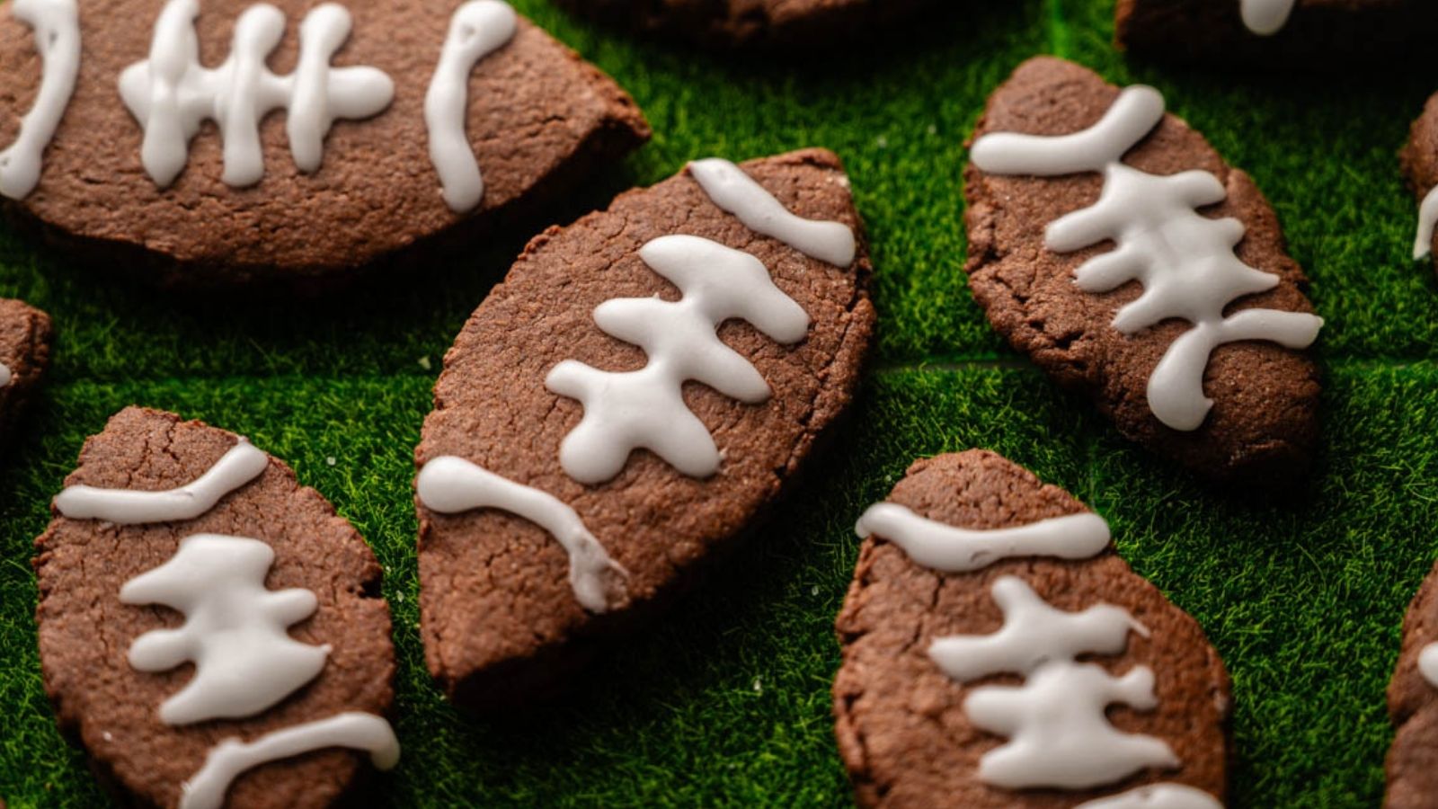 Football-shaped chocolate cookies with white icing laces are arranged on a green textured surface resembling grass.
