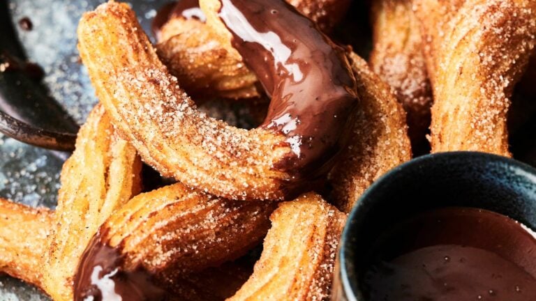 Close-up of churros coated in sugar, partially dipped in chocolate sauce, with a bowl of chocolate sauce nearby.
