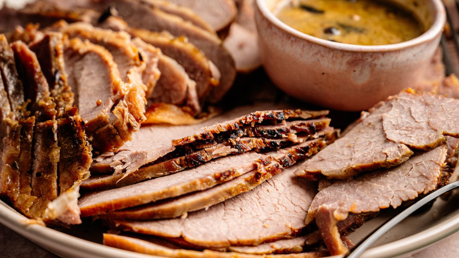 Sliced roast beef arranged on a plate with a bowl of gravy or sauce beside it.
