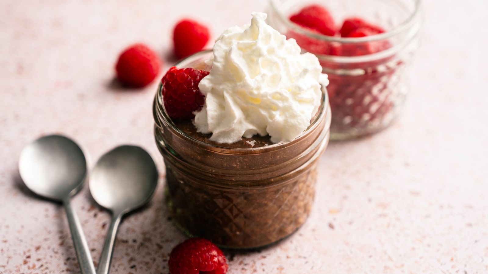 A small glass jar filled with chocolate mousse, topped with whipped cream and raspberries, sits beside two spoons and another jar with raspberries on a light surface.