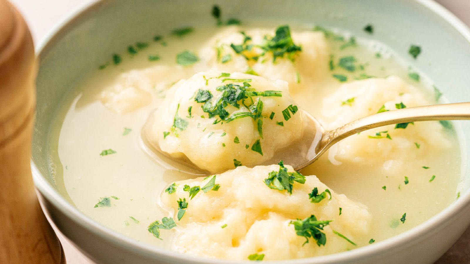 A bowl of dumpling soup with three dumplings in clear broth, garnished with chopped parsley, and a spoon lifting one dumpling.