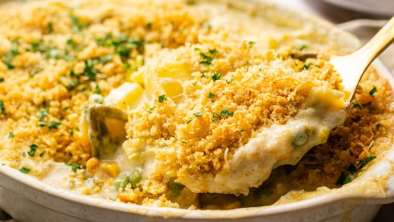 A close-up of a creamy vegetable casserole topped with golden breadcrumb crust, being scooped with a serving spoon.