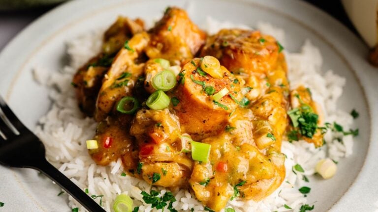 A plate of white rice topped with pieces of chicken in a yellow curry sauce, garnished with chopped green onions and herbs.