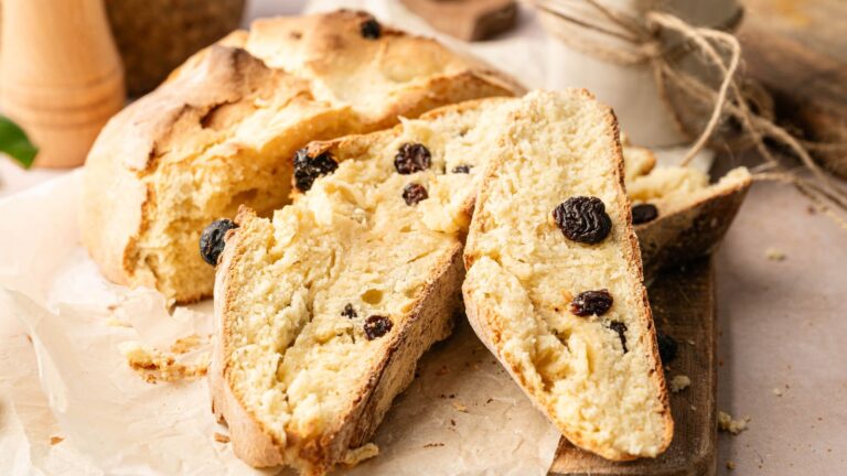 A loaf of Irish soda bread with raisins, partially sliced, rests on parchment paper on a wooden cutting board.