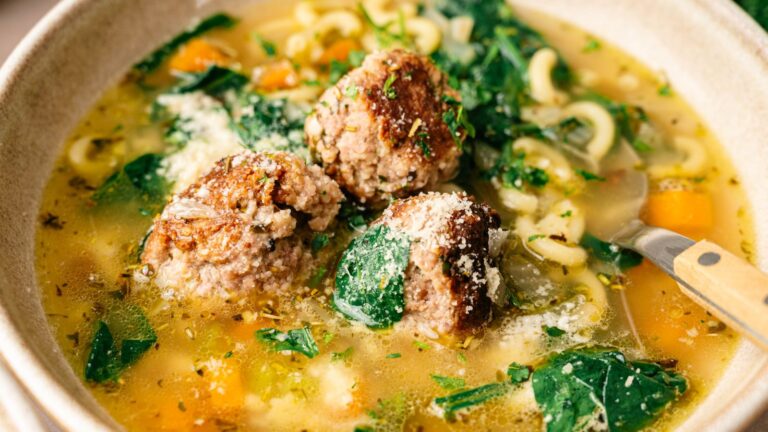 A bowl of soup with meatballs, pasta, spinach, diced vegetables, and broth, topped with grated cheese and herbs, with a spoon in the bowl.