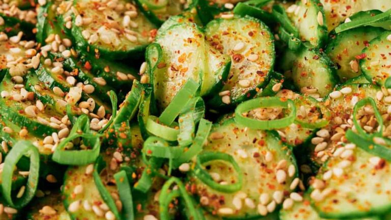 Sliced cucumbers topped with sesame seeds, chopped green onions, and red pepper flakes, shown in a close-up view.