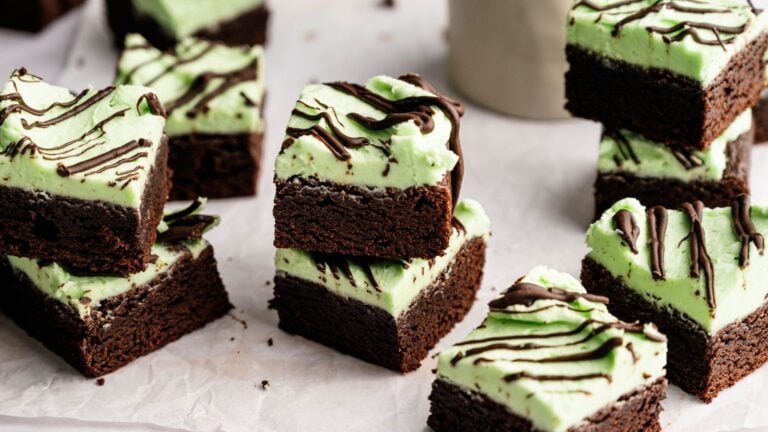 Several mint chocolate brownies with a green mint frosting layer and chocolate drizzle are stacked and arranged on parchment paper.
