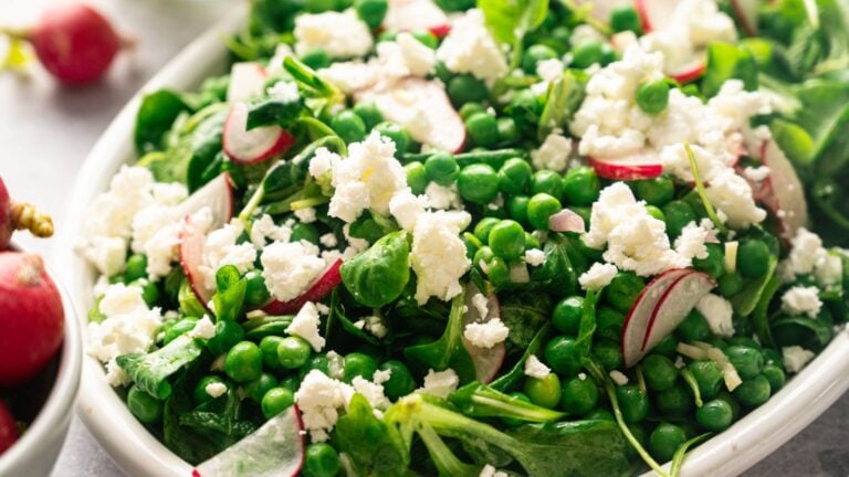 A close-up of a fresh salad with leafy greens, sliced radishes, green peas, and crumbled white cheese on a white plate.