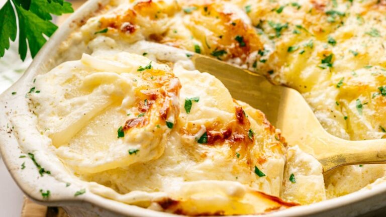 A close-up of creamy, baked scalloped potatoes topped with melted cheese and sprinkled with fresh herbs, being served with a golden spatula.