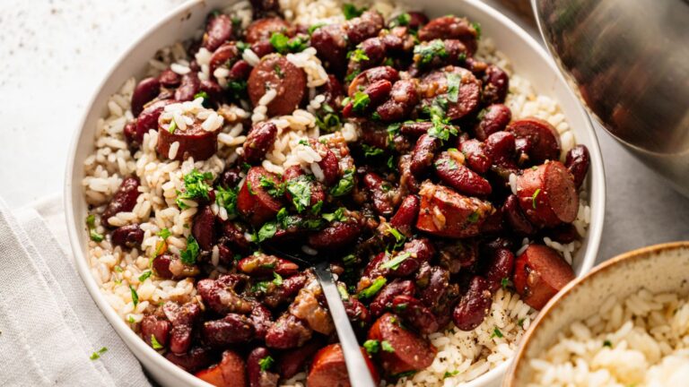A bowl of white rice topped with red beans, sliced sausage, and chopped herbs, with a fork resting in the bowl.