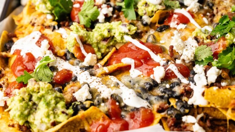 A close-up of loaded nachos topped with melted cheese, black beans, ground meat, guacamole, sour cream, salsa, crumbled cheese, and cilantro.