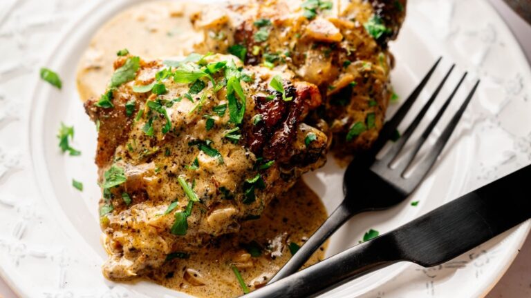 Cooked chicken in a creamy sauce garnished with chopped herbs, served on a white plate with a black fork and knife.
