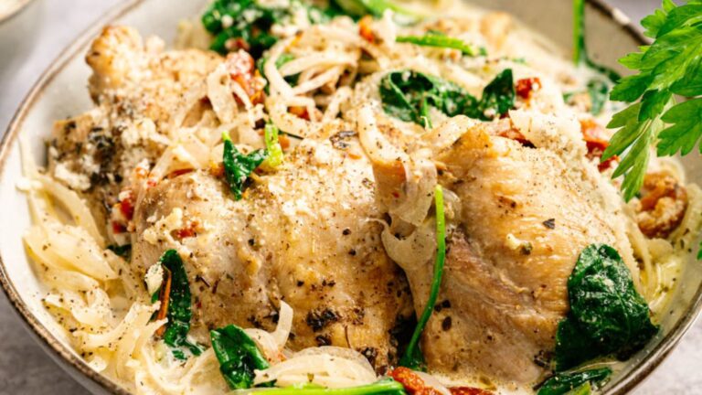 Creamy chicken thighs served with sautéed onions, spinach, and sun-dried tomatoes, garnished with ground black pepper and herbs in a bowl.