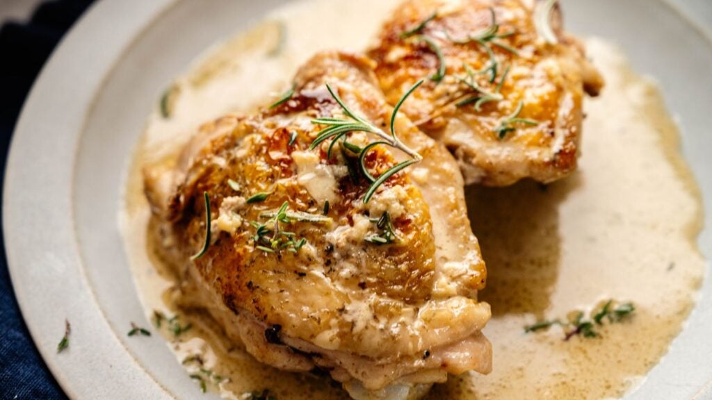 Two pieces of roasted chicken with crispy skin are served on a plate with creamy sauce and garnished with fresh herbs.
